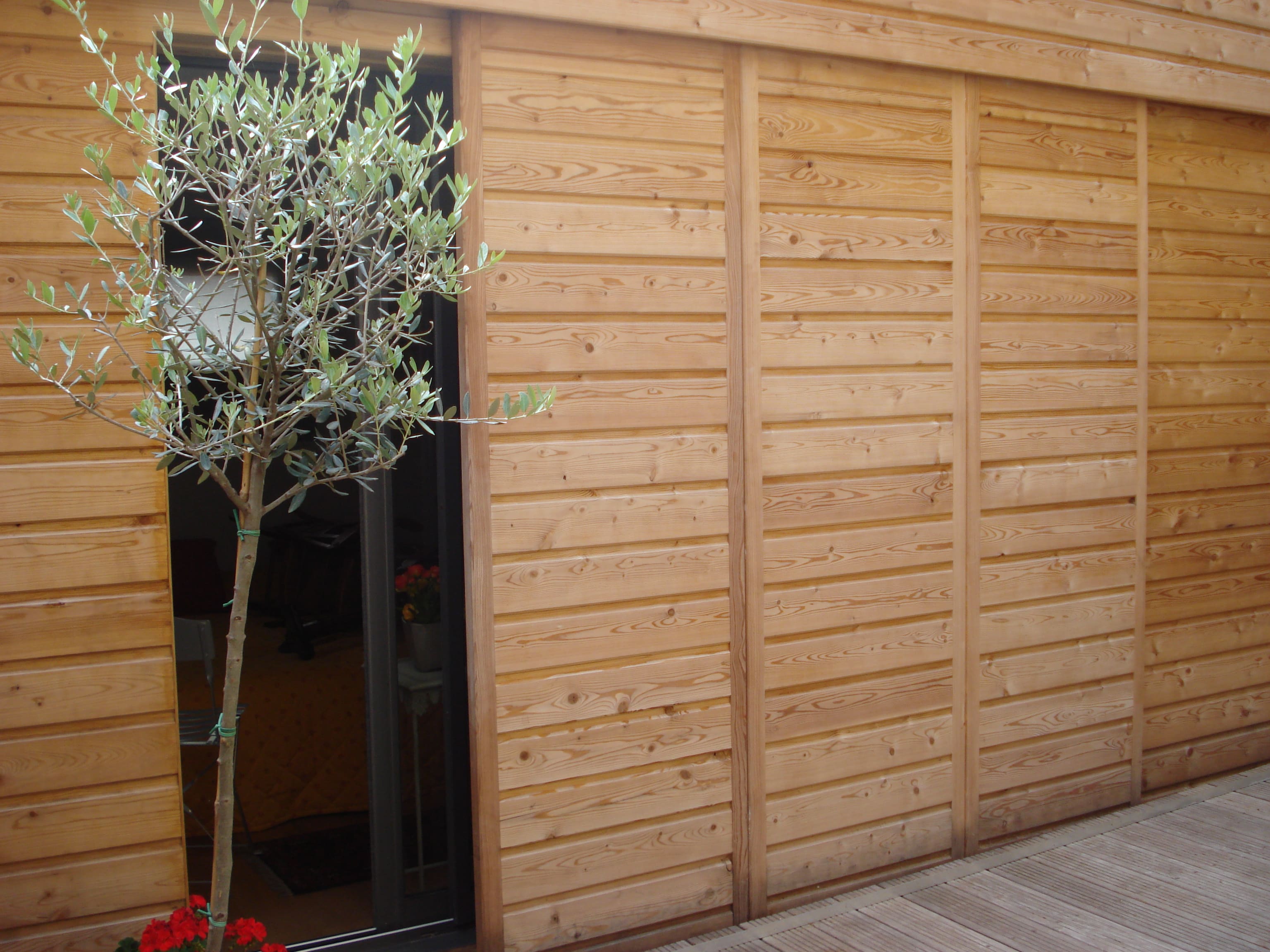 Patio d'appartement habillé d'un bardage bois horizontal aux grands panneaux coulissants, olivier en pot et terrasse en bois