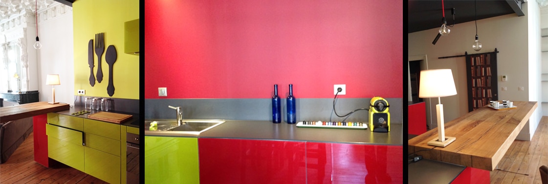 Détails de la cuisine : meubles vert anis et couverts géants au mur, mur rouge au plan de travail gris, table-bar en chêne sous suspensions à cordon rouge et porte coulissante de bibliothèque