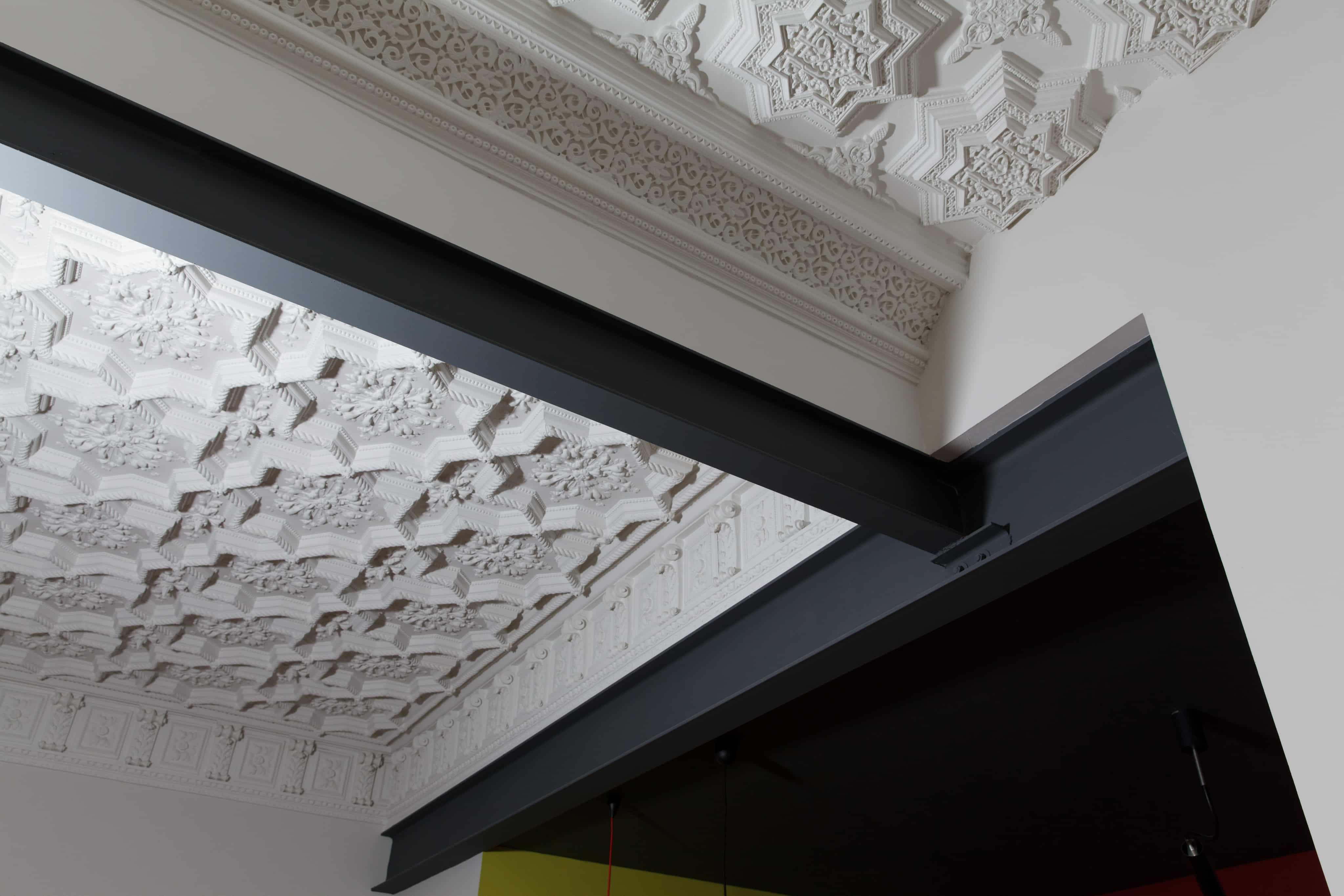 Plafond ancien aux plâtres moulurés traversé par une poutre d'acier noir, contraste entre ornements blancs et ligne contemporaine