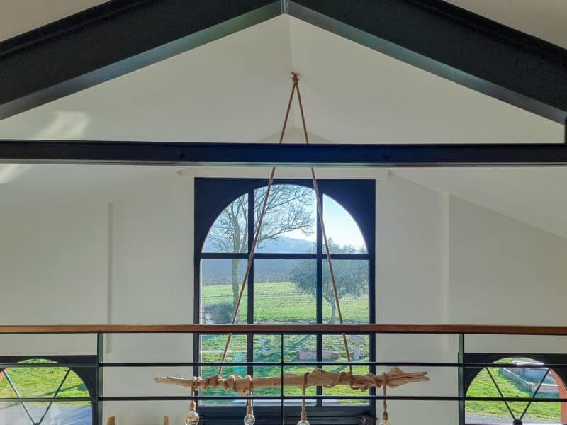 Mezzanine sous charpente noire face à une grande baie cintrée ouverte sur la campagne, luminaire en bois flotté suspendu