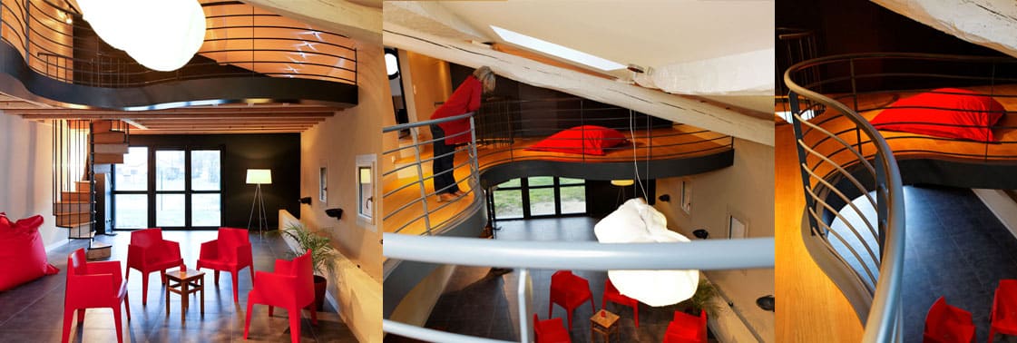 Espace polyvalent aux fauteuils rouges et poufs, mezzanine courbe desservie par un escalier hélicoïdal sous une grande suspension nuage
