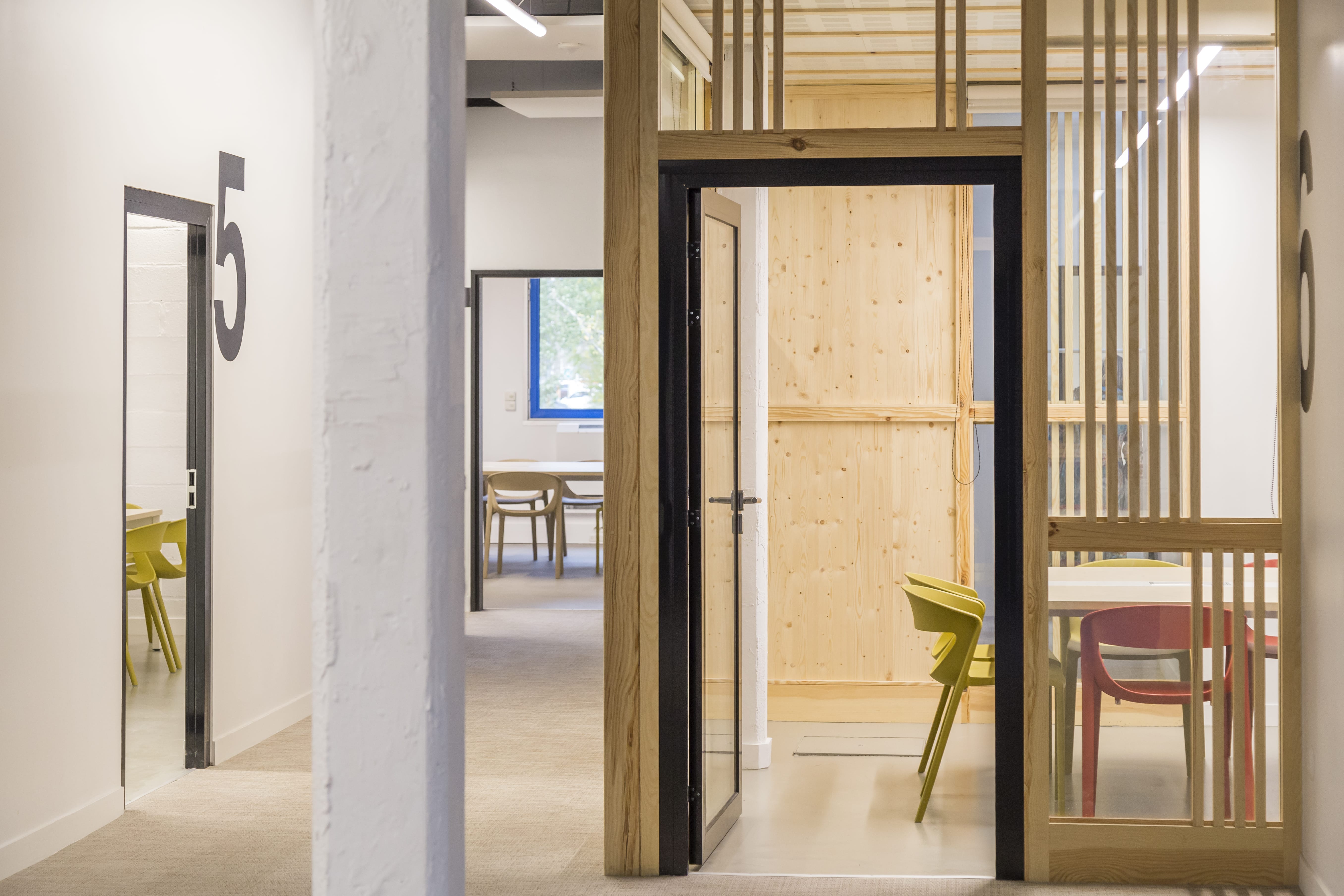 Business center Le 457 : couloir aux salles de réunion numérotées, cloisons en contreplaqué de pin et claustras bois, chaises colorées jaune et rouge
