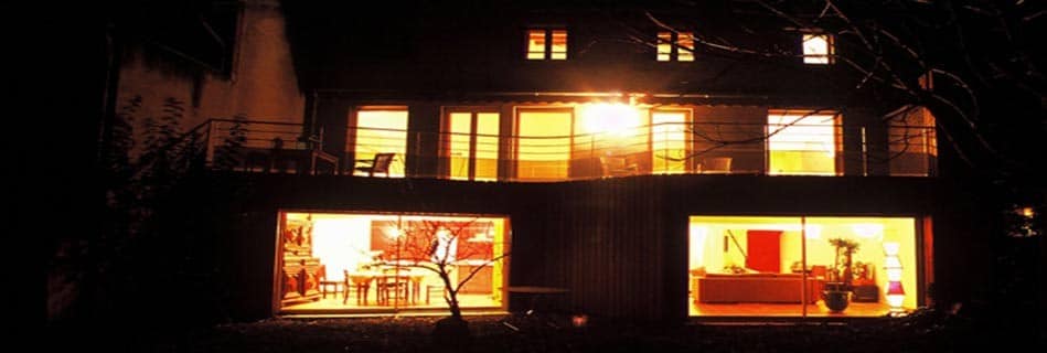 Façade jardin la nuit : la double hauteur du séjour éclairée derrière ses grandes baies, surmontée de la terrasse bois et de son garde-corps courbe