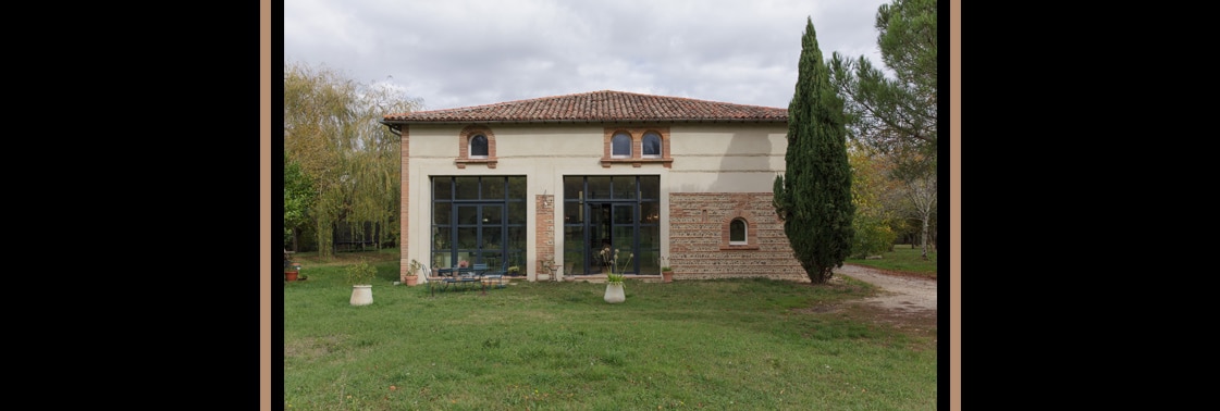 La ferme réhabilitée aux enduits clairs et toiture en tuile canal à quatre pentes, pignon de brique, hautes baies vitrées à ossature d'acier et cyprès dans le jardin