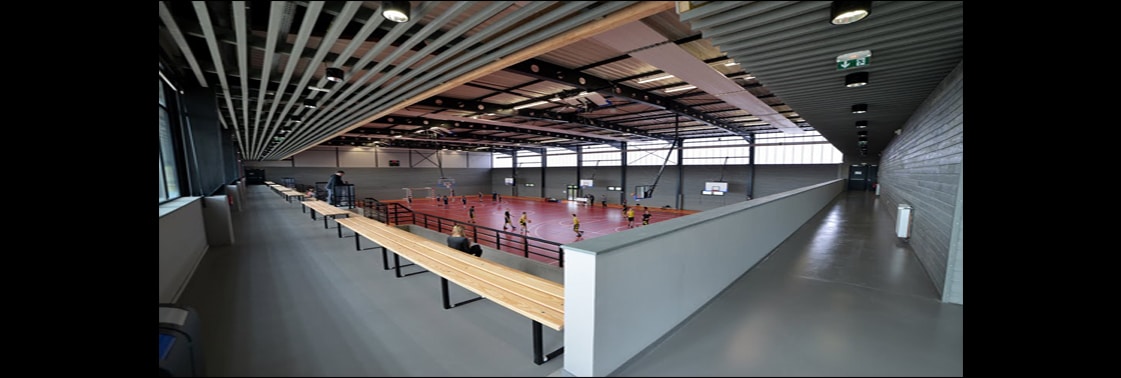 Coursive et gradins en bois surplombant le terrain rouge du gymnase, sous un plafond à résilles bois et métal