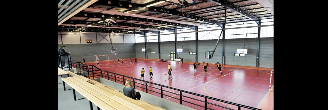 Match sur le terrain rouge du gymnase, paniers et buts en place sous la charpente métallique et les verrières hautes