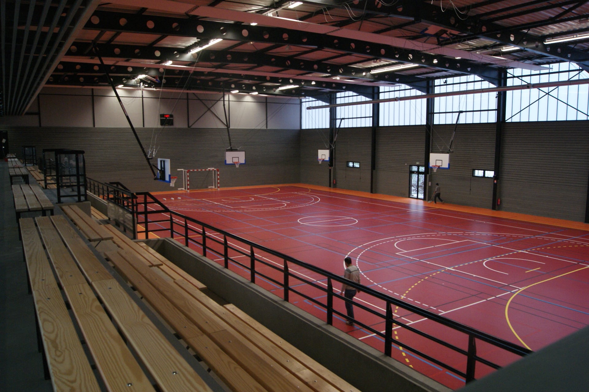 Grande salle du gymnase vue depuis les gradins en bois : terrain rouge tracé pour plusieurs sports, paniers de basket et charpente métallique sous verrières hautes