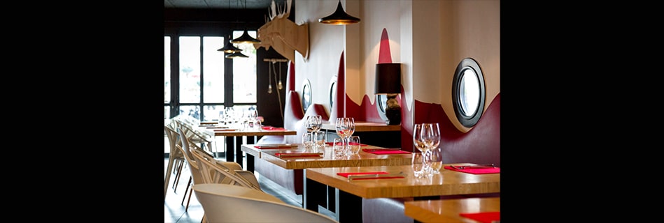 Salle du restaurant : trophée de cerf en carton, banquettes rouges en silhouette de montagne et tables en bois dressées de rouge