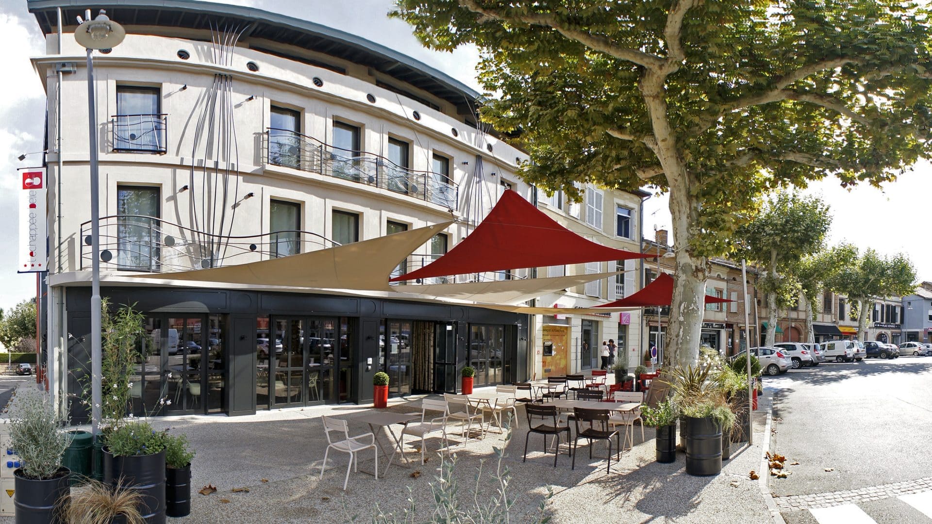 Hôtel L'Échappée Belle : façade blanche courbe aux balcons en ferronnerie à motifs de pissenlit, restaurant en rez-de-chaussée et terrasse ombragée de voiles sous les platanes