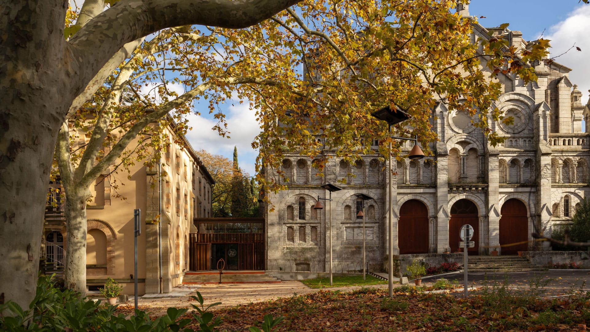 Ensemble monastique dominicain : la grande église romane en pierre et le couvent en enduit ocre, reliés par un nouveau pavillon d'entrée en bois et verre, sous un platane d'automne
