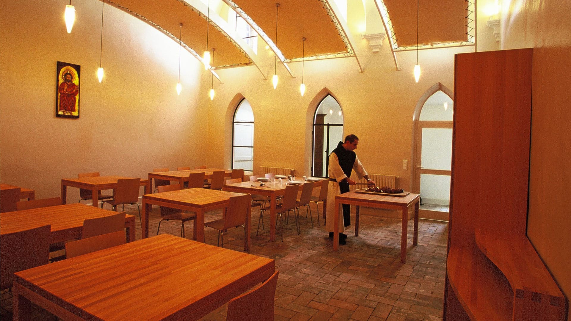 Réfectoire voûté de l'hôtellerie du monastère : tables et chaises en bois clair sur sol de tomettes, fenêtres gothiques et moine dressant le service, sous une icône du Christ