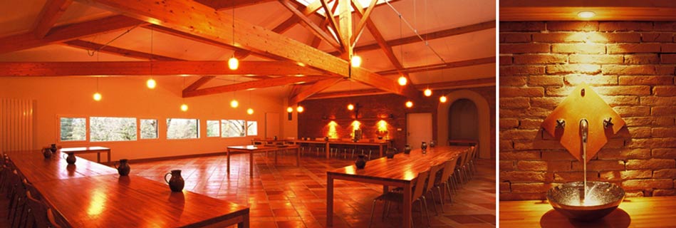 Réfectoire aux longues tables de bois et pichets en terre sous la charpente, et détail d'un lavabo en pierre à bec métallique contre un mur de brique