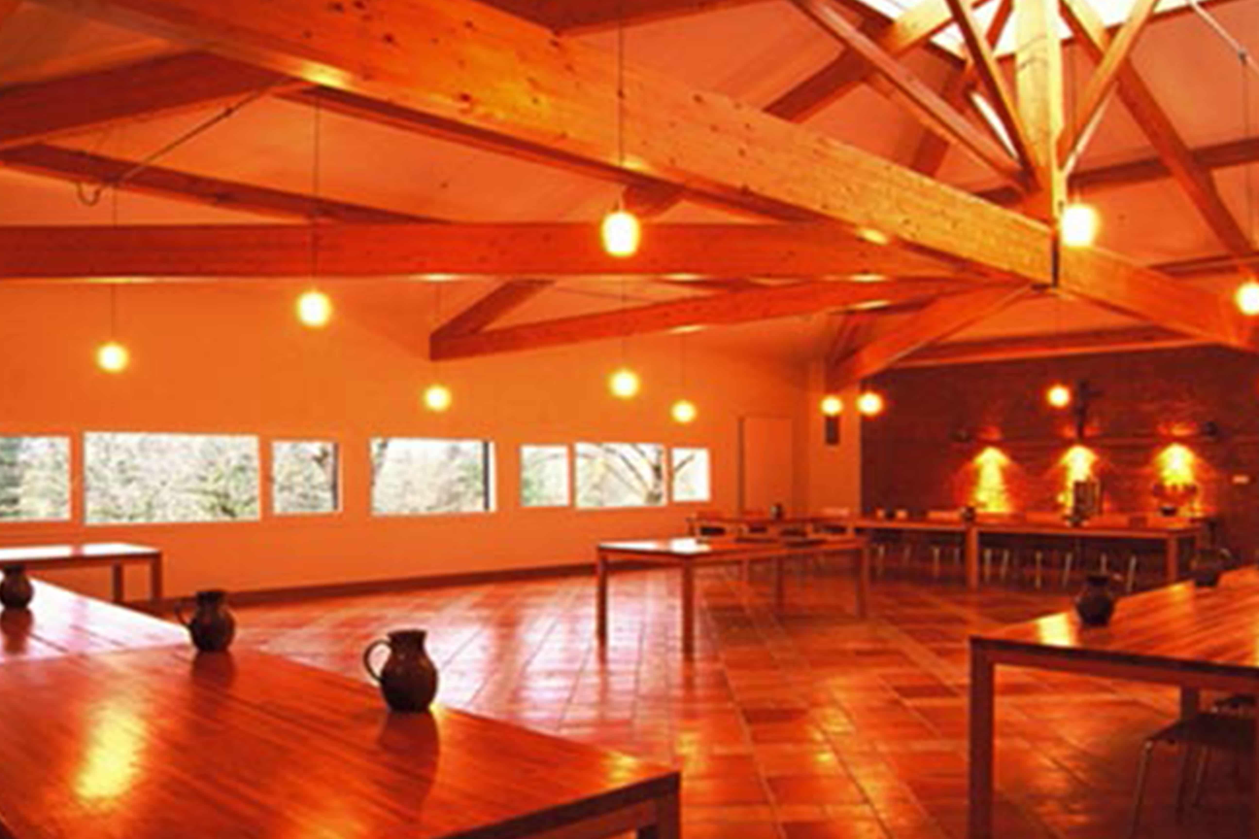 Vaste réfectoire d'abbaye : longues tables en bois et pichets en terre, sol en tomettes et charpente apparente sous une lumière chaude, ruban de fenêtres sur la campagne