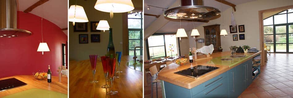 Cuisine ouverte sous charpente apparente : îlot central bleu-vert au plan de travail bois et verre, hotte ronde en inox, mur framboise, table en bois et grande baie vitrée sur le jardin