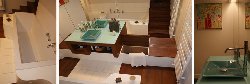 Salle de bains en verre turquoise et bois foncé : baignoire au tablier arrondi, meuble vasque au plan de verre translucide et deux vasques, marches en bois et tableau coloré