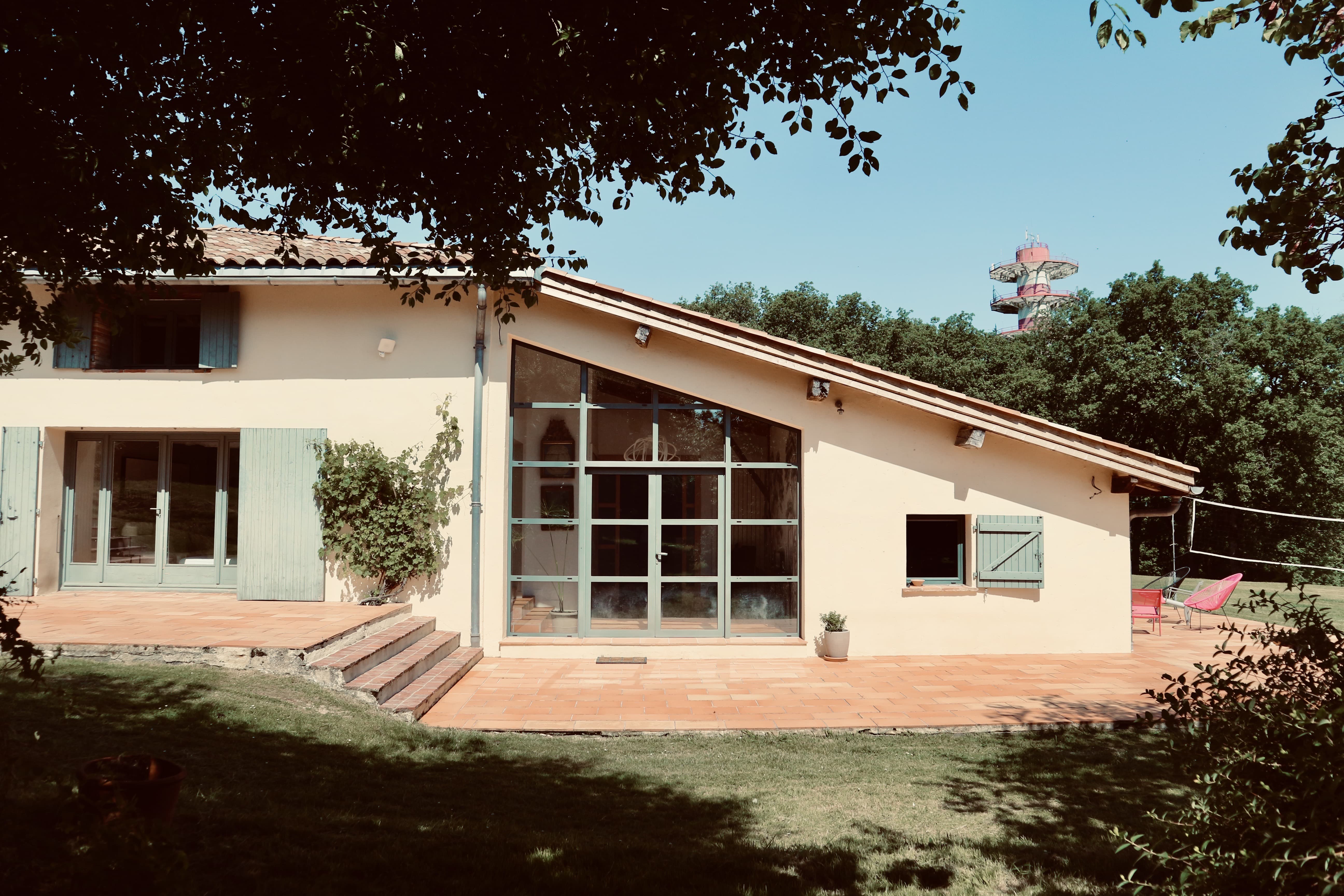Maison de campagne aux murs crème et volets vert pâle, prolongée par une extension en appentis à grande verrière ouvrant sur une terrasse en tomettes et le jardin