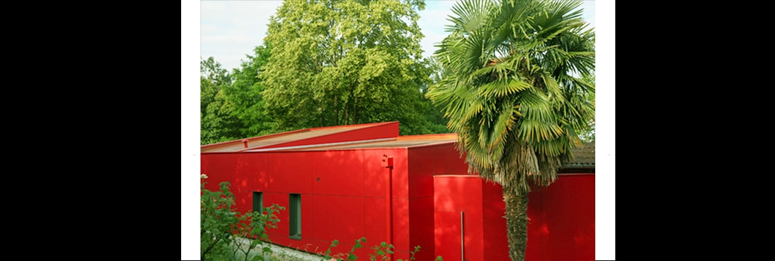 L'extension aux façades en résine composite rouge et pans coupés, avec sa toiture cuivrée, au pied d'un palmier dans le jardin arboré