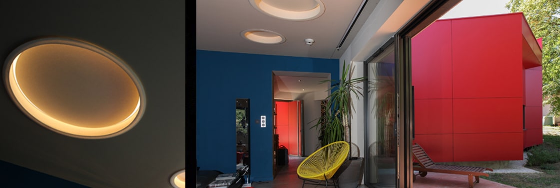 Entrée aux murs bleu profond et plafonds circulaires rétroéclairés, ouverte par une baie vitrée sur le patio et les volumes rouges de l'extension