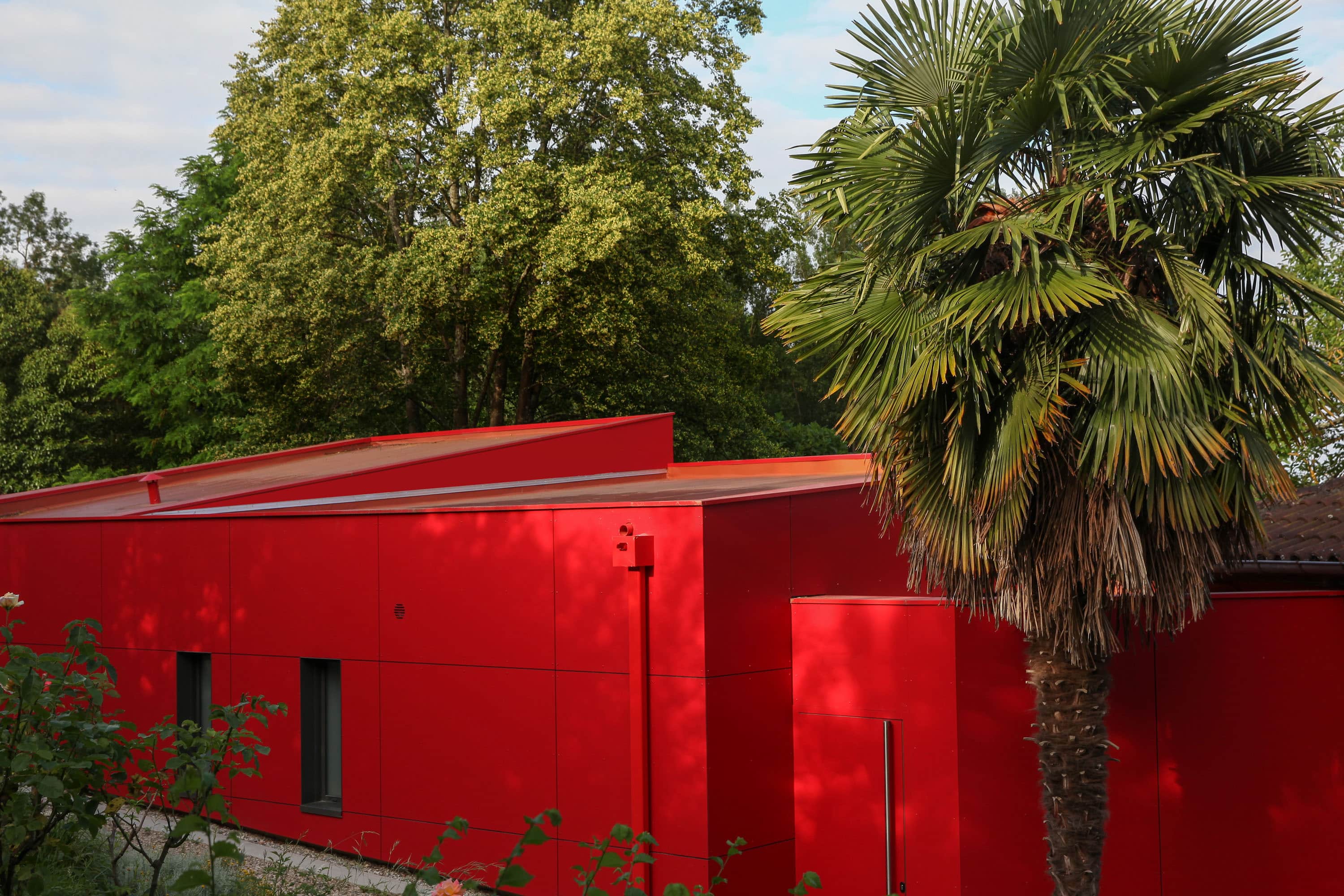 Extension contemporaine aux façades en résine composite rouge et pans coupés en focale, au pied d'un palmier et d'arbres verts dans un jardin arboré