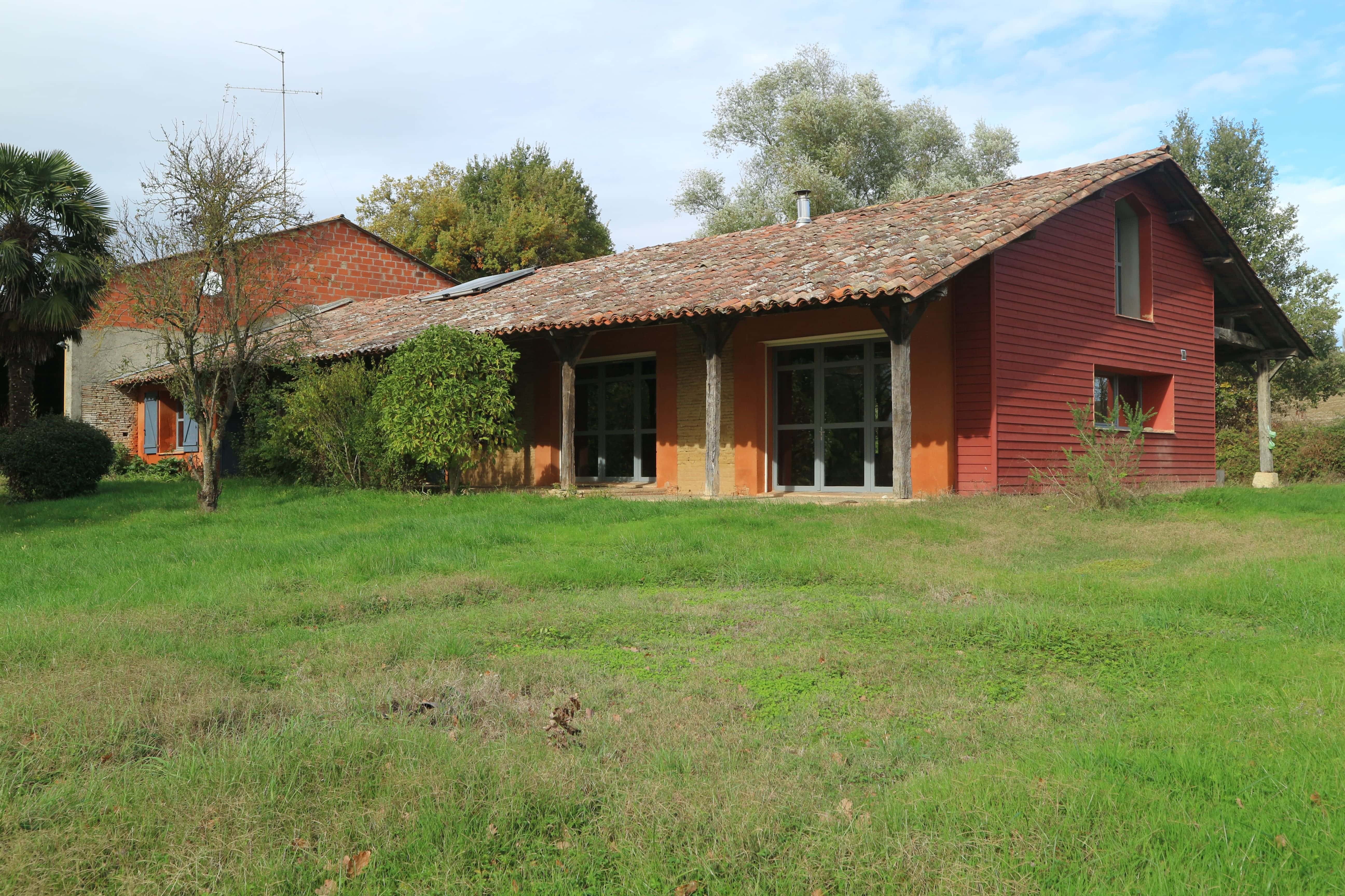 Ferme en terre aux murs enduits d'argile ocre, longs auvents débordants et pignon en bardage bois peint rouge, posée dans une prairie