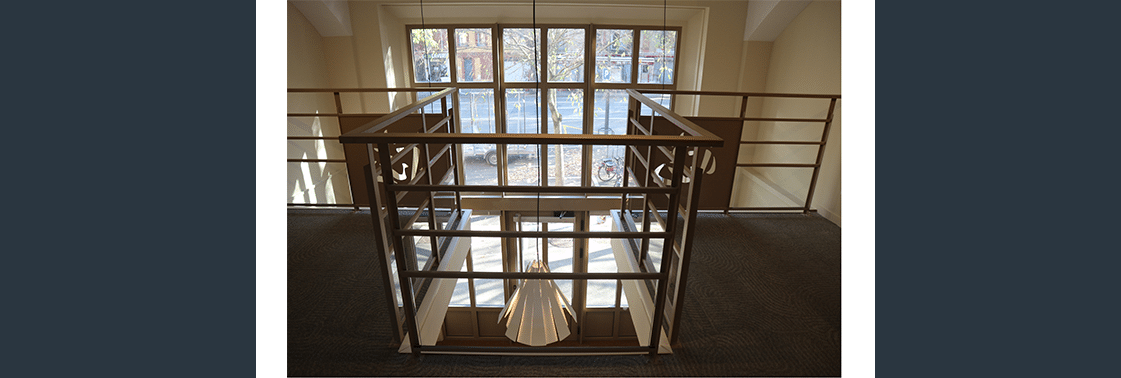 Espace en double hauteur : mezzanine à garde-corps bois surplombant de hautes fenêtres sur l'avenue plantée, sol moquette et suspension plissée