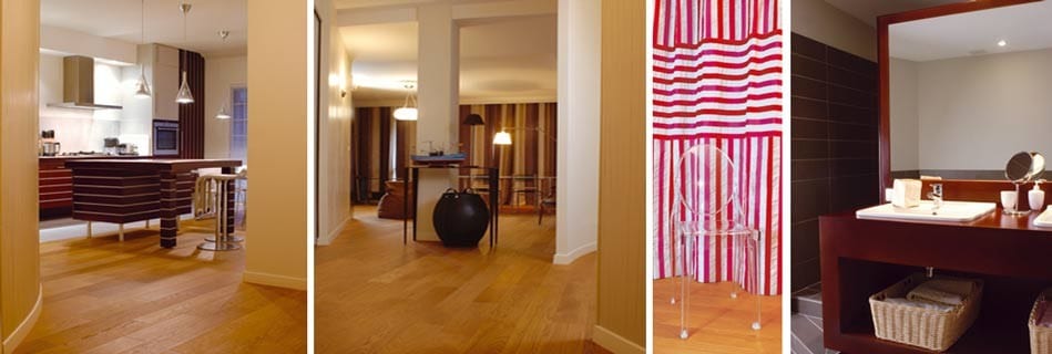 Enfilade de l'appartement sur parquet de chêne : cuisine ouverte, séjour et couloir, chaise transparente devant un rideau rayé et salle de bains habillée de bois sombre
