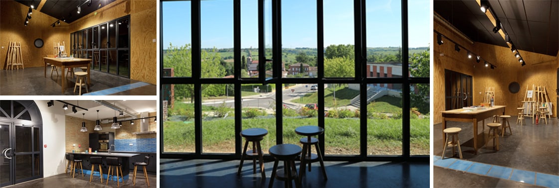 Salle d'activités aux murs en panneaux OSB avec chevalets de peinture, coin cuisine et bar, et grande verrière d'acier noir ouverte sur le paysage vallonné