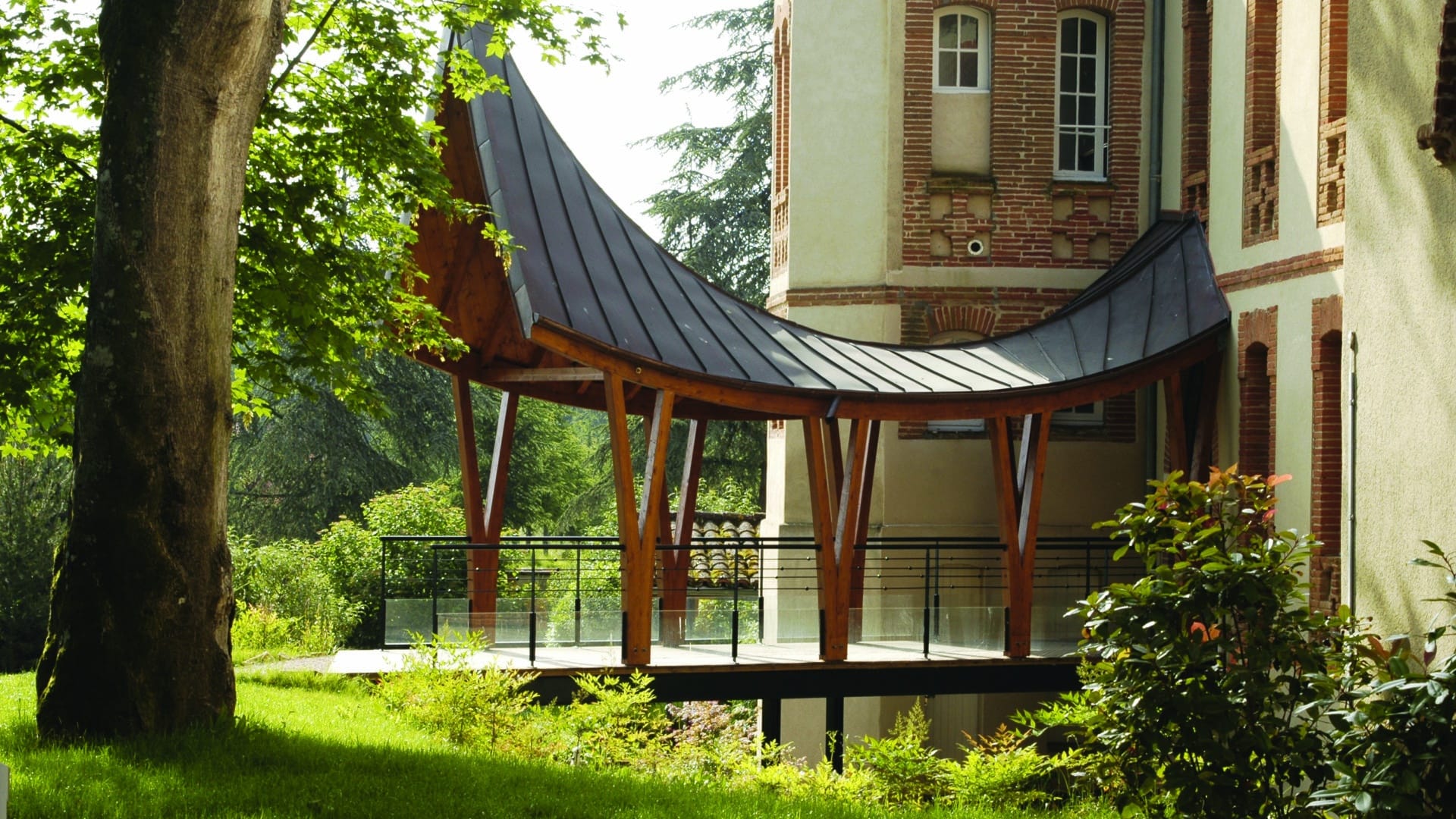 Nouvel auvent d'accès au monastère : une toiture de zinc en courbe posée sur des poteaux bois, reliant le jardin au bâtiment ancien en brique