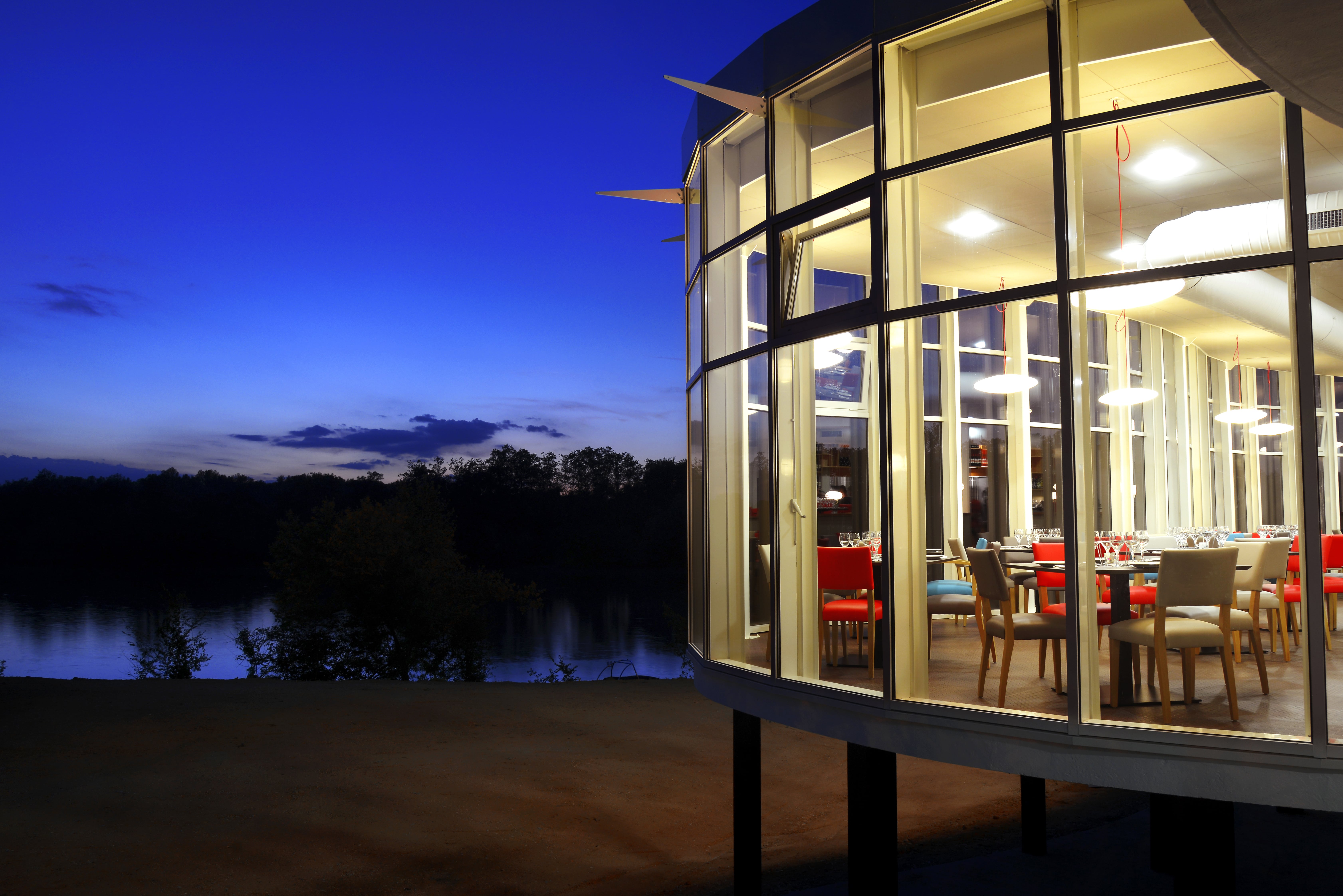 Le restaurant Le Clapotis au crépuscule : sa rotonde vitrée éclairée en surplomb de la rivière, salle garnie de chaises rouges et crème sous un ciel bleu nuit