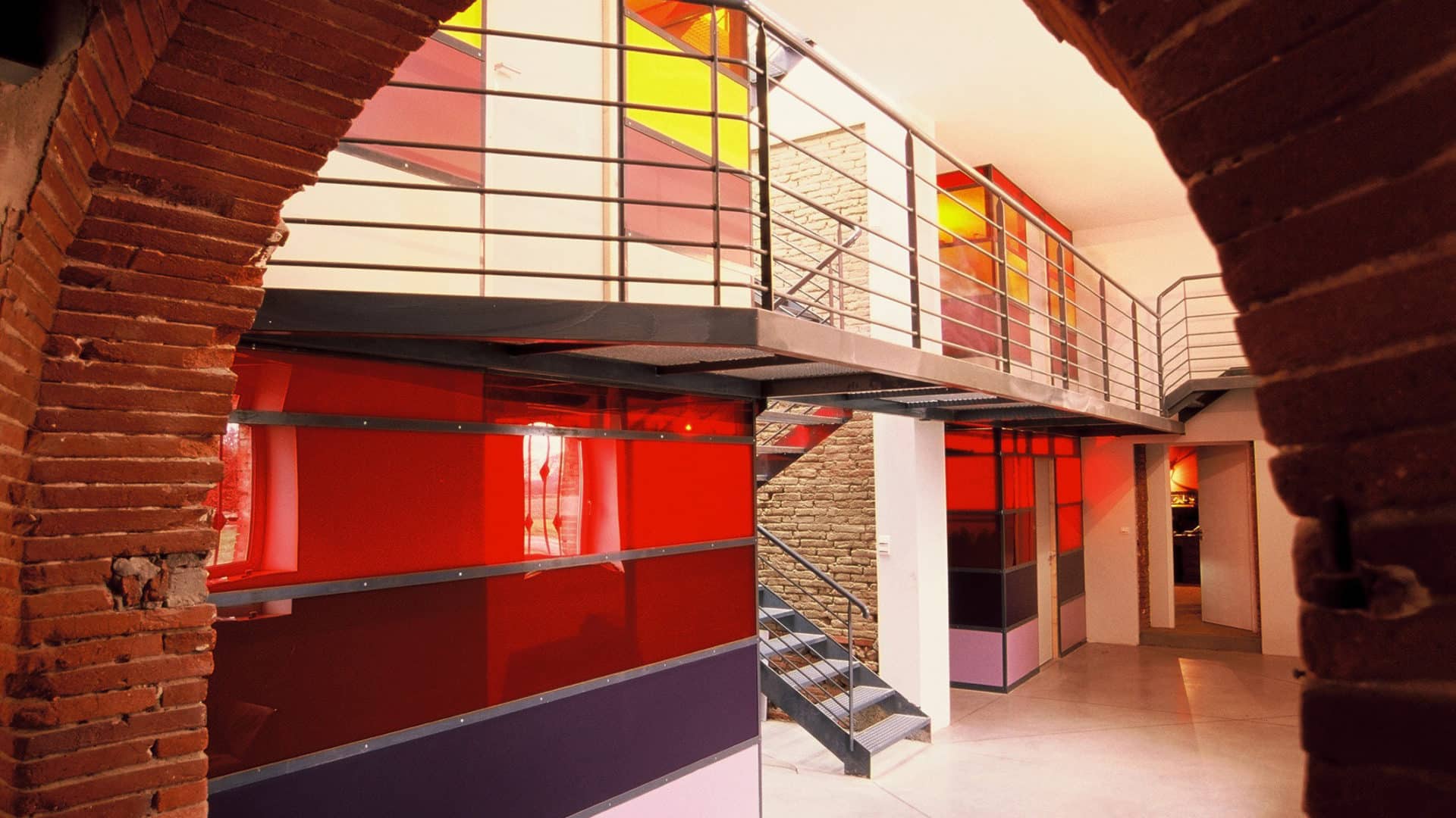 Mezzanine aux blocs de verre coloré rouge, orange et jaune, reliés par une passerelle en caillebotis métallique, vue sous une arche de brique