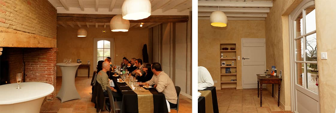 Salle de séminaire à la grande cheminée de brique où déjeune un groupe sous des suspensions en papier, et entrée aux niches-bibliothèque près d'une fenêtre cintrée