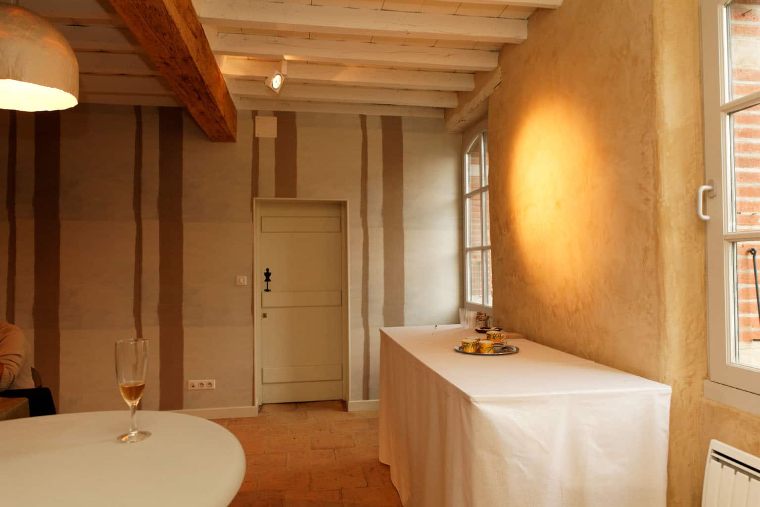 Salle de réception aux poutres apparentes et murs rayés, sol en tomettes, buffet dressé et coupe de champagne près d'une fenêtre cintrée
