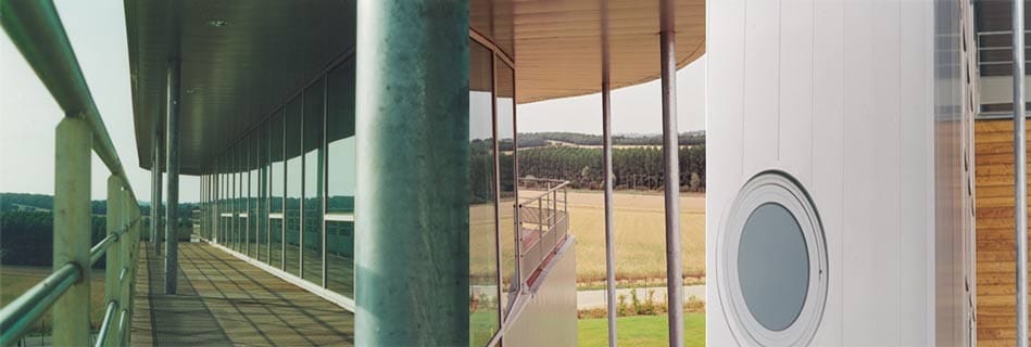 Galerie périphérique vitrée à ossature d'acier et platelage bois ouverte sur la campagne, et détail d'un oculus rond sur le bardage blanc