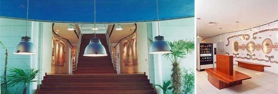 Hall d'accueil au grand escalier de bois sous des suspensions industrielles et fresques en mosaïque, et banque d'accueil devant une mosaïque au motif de globe