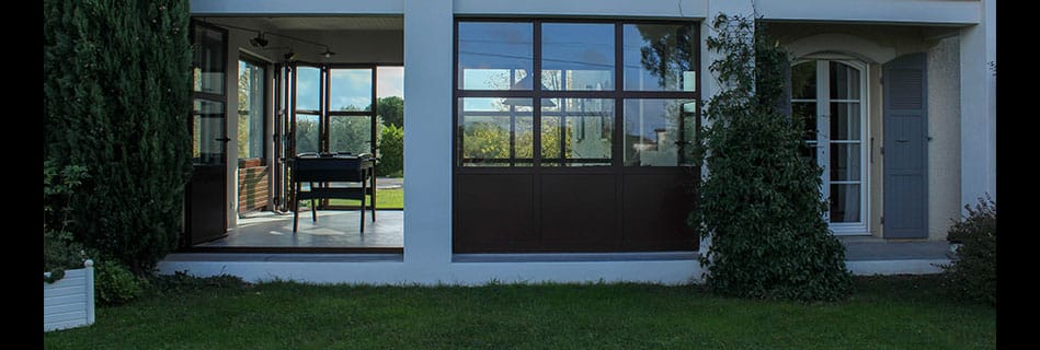 Façade de la nouvelle pièce vitrée au crépu blanc, menuiseries bois pliantes ouvertes sur la terrasse, vue depuis le jardin au crépuscule